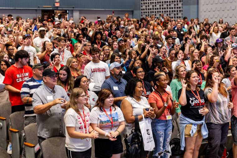 A large group of students and parents attending Orientation stand and clap in an auditorium 
