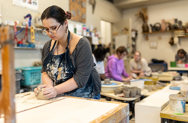UL Lafayette ceramics student working with clay at a bench with classmates behind working at their own stations.