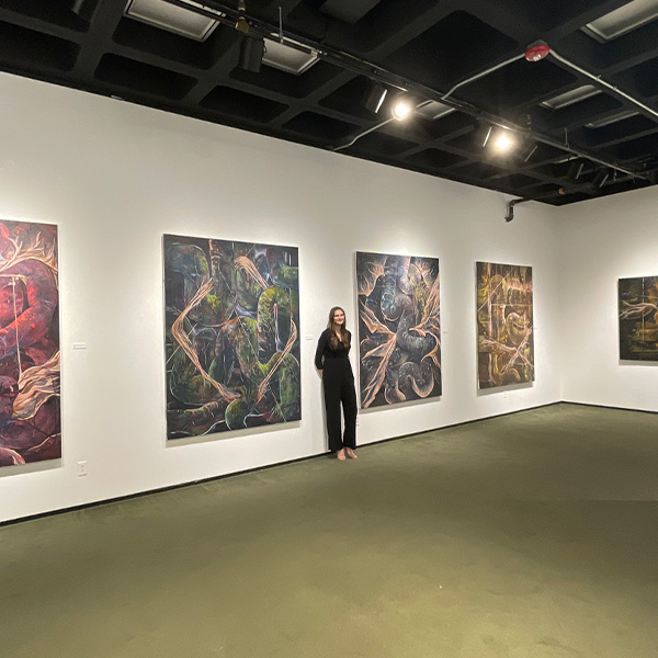 UL Lafayette painting student standing in front of a gallery wall of painted canvases.