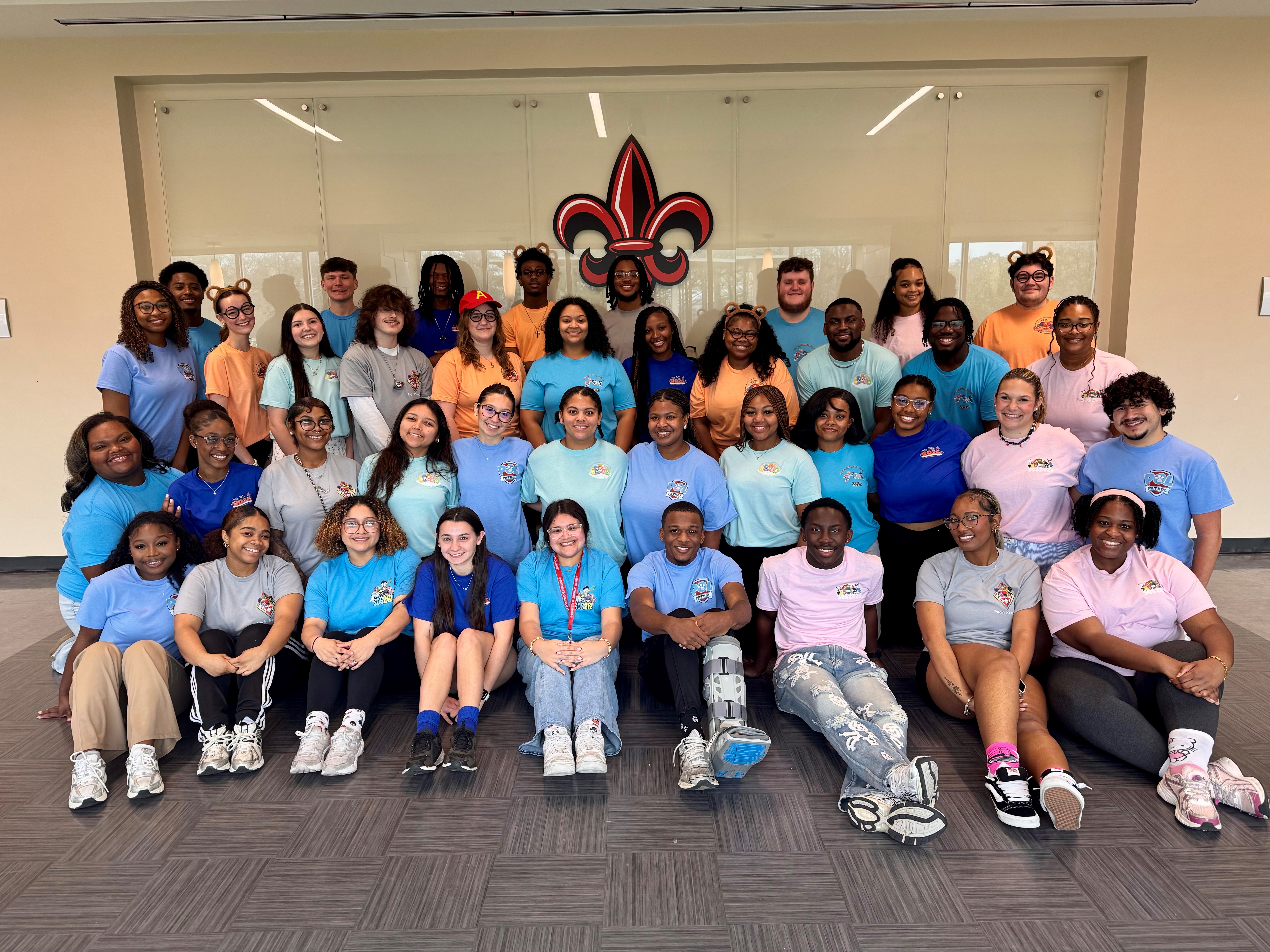 Group photo of the 2026 Student Orientation Staff in front of a white wall with a fleur de lis on the wall. 