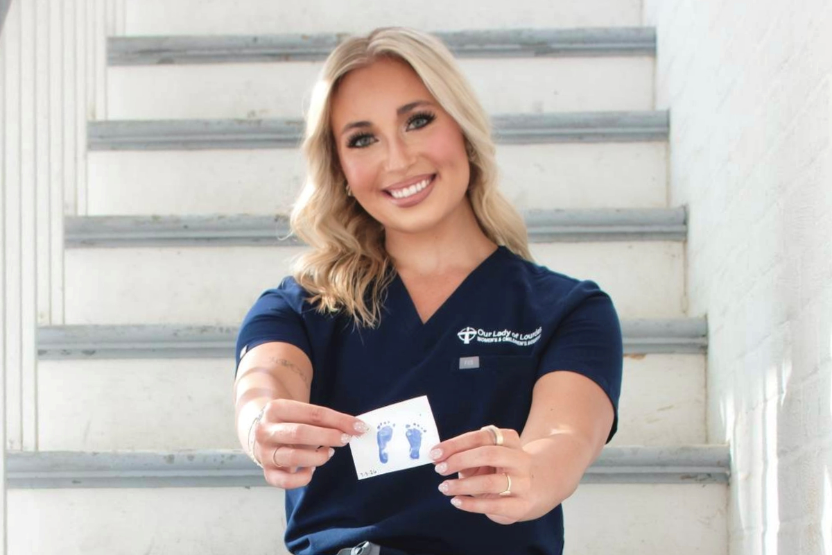 Graduate of UL Lafayette's online RN to BSN sits on a staircase wearing scrubs, holding a NICU emblem.