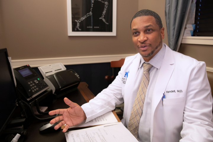 Tim Randell, M.D. works as an orthopedic surgeon at his desk wearing a white coat and beige tie.