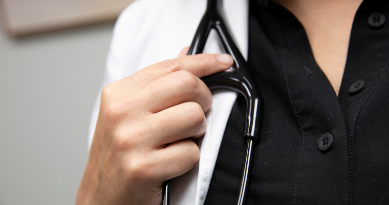 Online nursing student holds her stethoscope wearing black scrubs and a white coat.