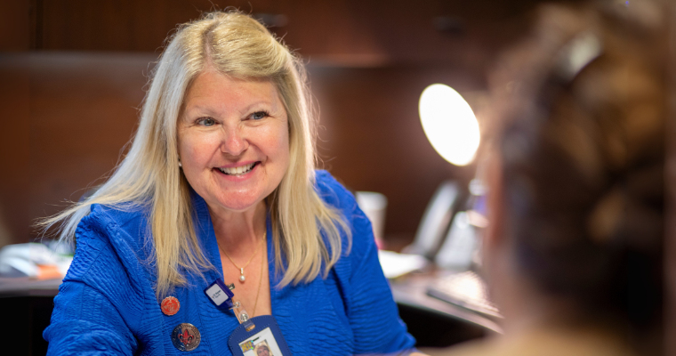 Online healthcare degree graduate smiles across desk wearing a blue blazer and UL Lafayette Alumni pins. 