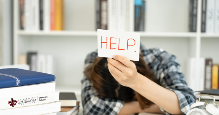 college student with head down at desk holding a sign in his hand that reads help