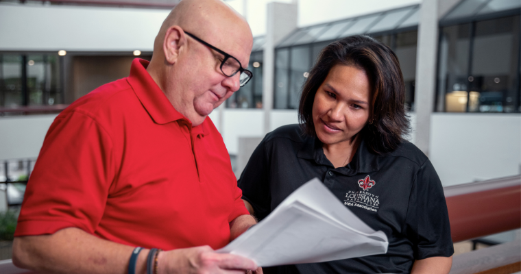 Online MBA graduate overviews paperwork with a colleague, wearing a black UL Lafayette MBA Assoc. polo.